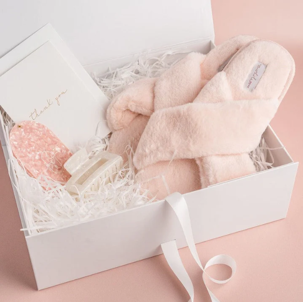 Pink fuzzy slippers inside a white gift box with shredded paper, a thank you card, and decorative soap or lotion bottles.
