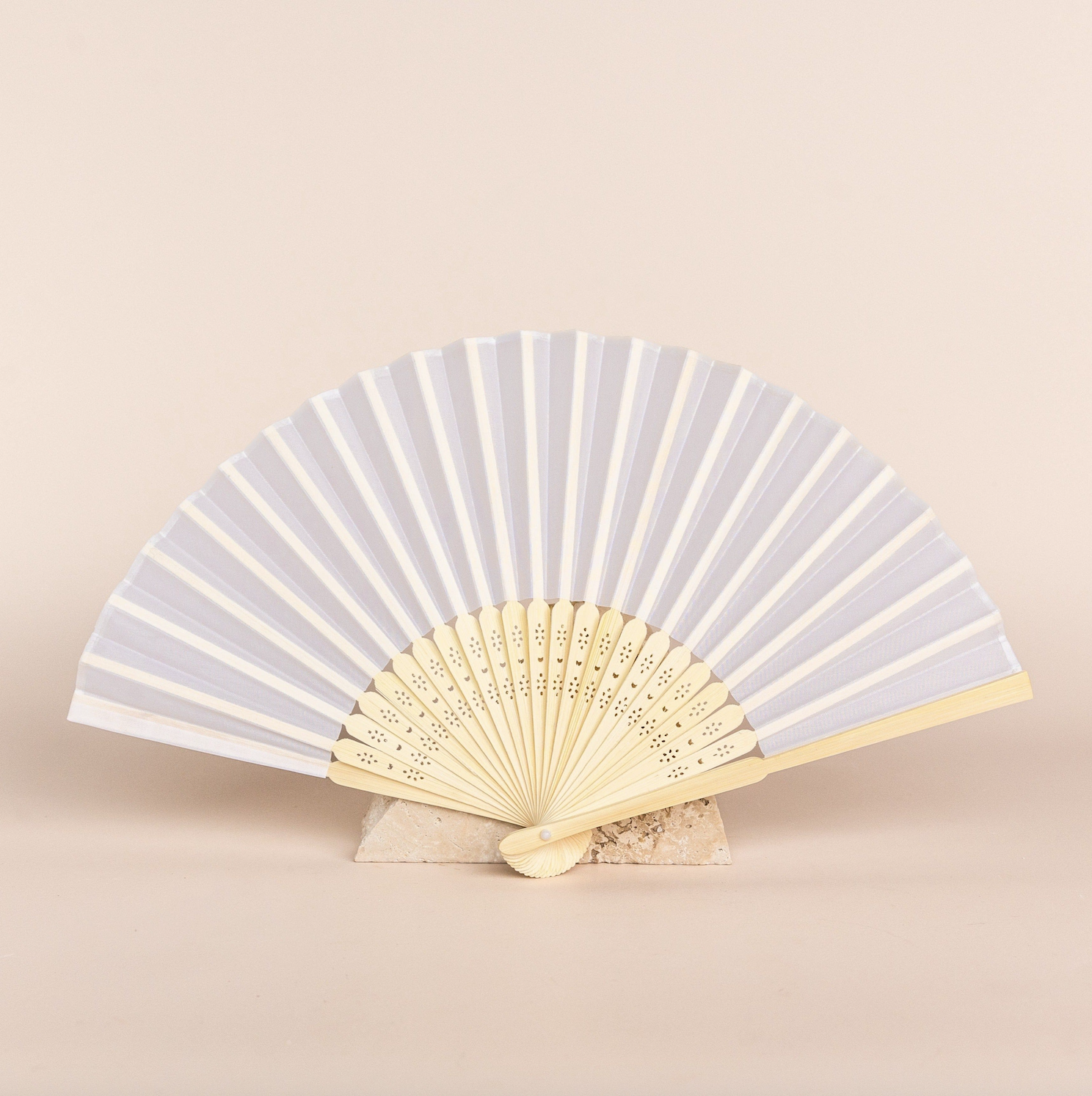A white folding hand fan with wooden ribs resting on a small stone block against a neutral background.