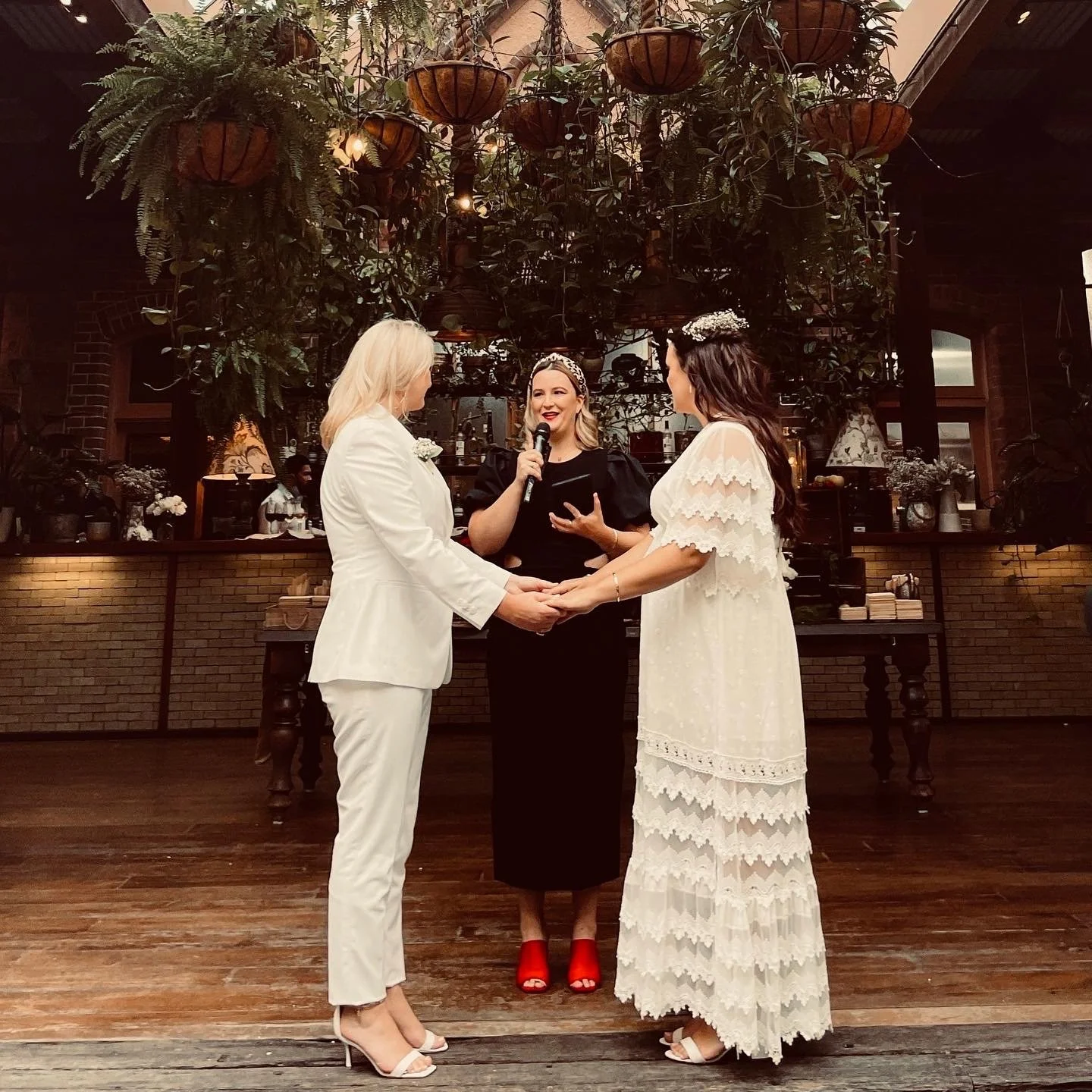 Two brides stand holding hands as the celebrant Married By Leah reads out the Monitum to them during their ceremony at The Grounds of Alexandria