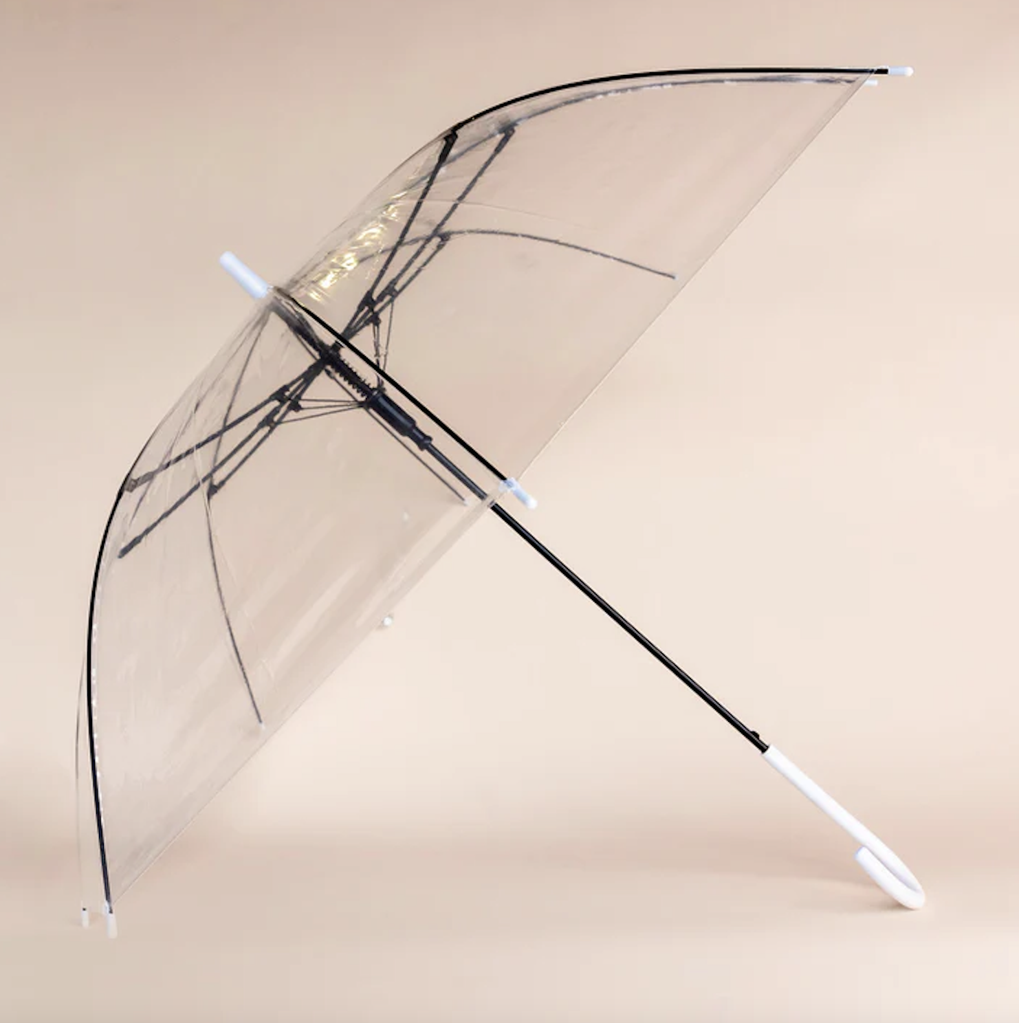 A transparent umbrella with black frame and white handle, open and leaning against a light beige background.
