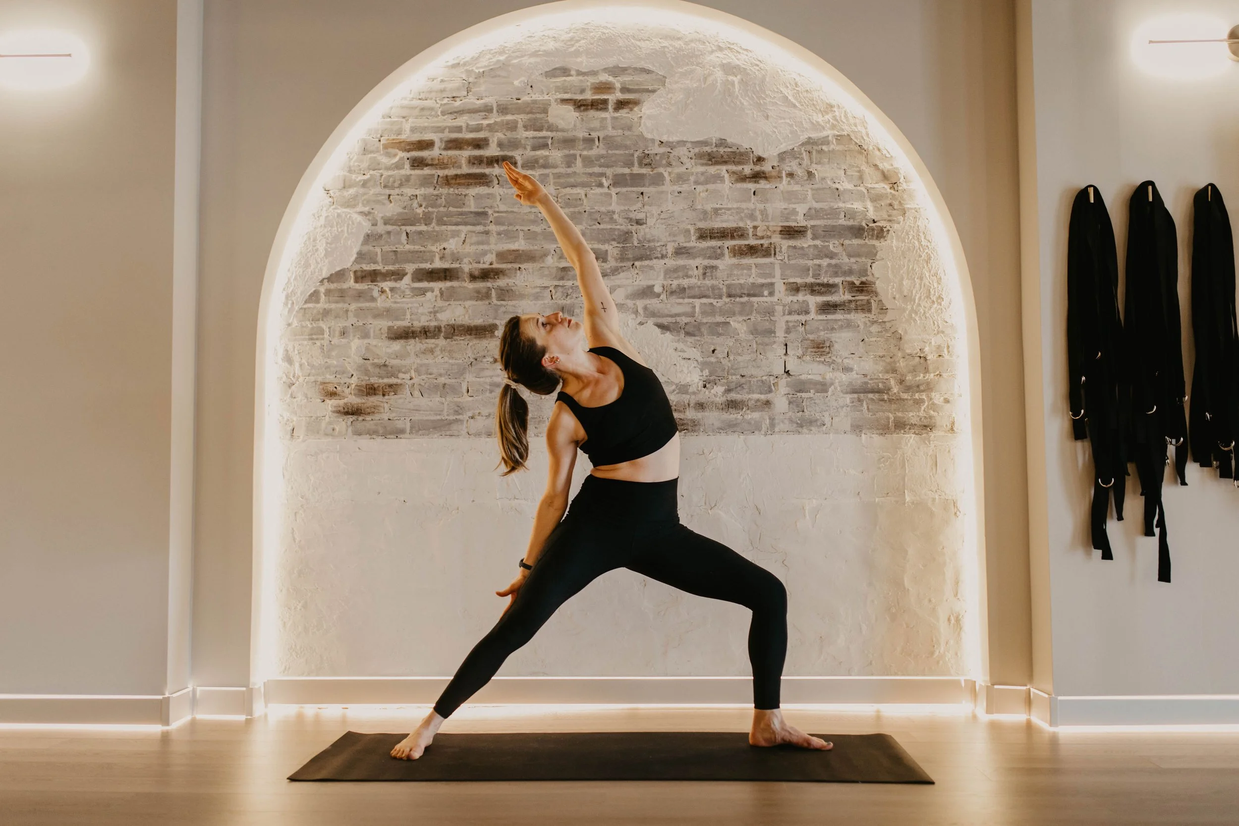 Woman practicing yoga in a modern studio with a brick and white wall background, wearing black athletic clothing, on a black yoga mat.