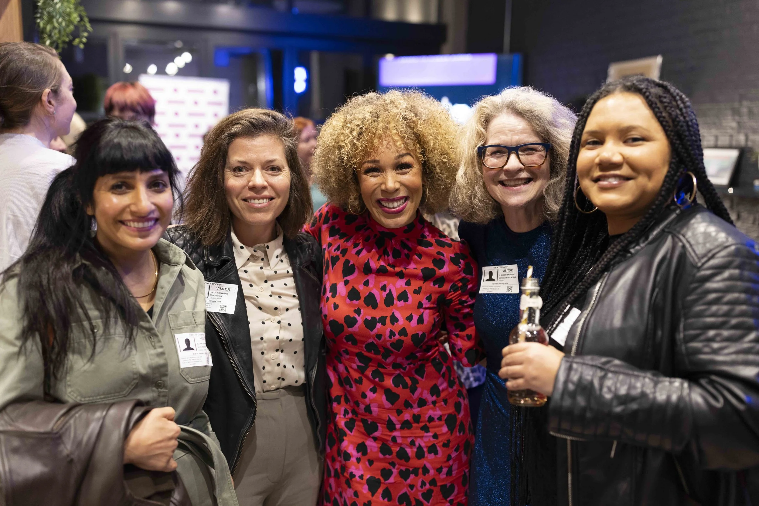 Group of five women smiling at an event, wearing name tags, with one holding a bottle.