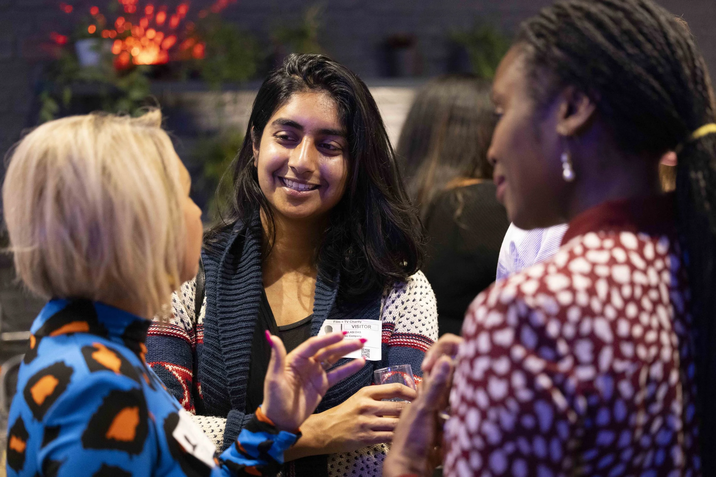 Three women engaged in conversation at a social event, with one woman smiling and wearing a patterned sweater, and the others listening attentively.