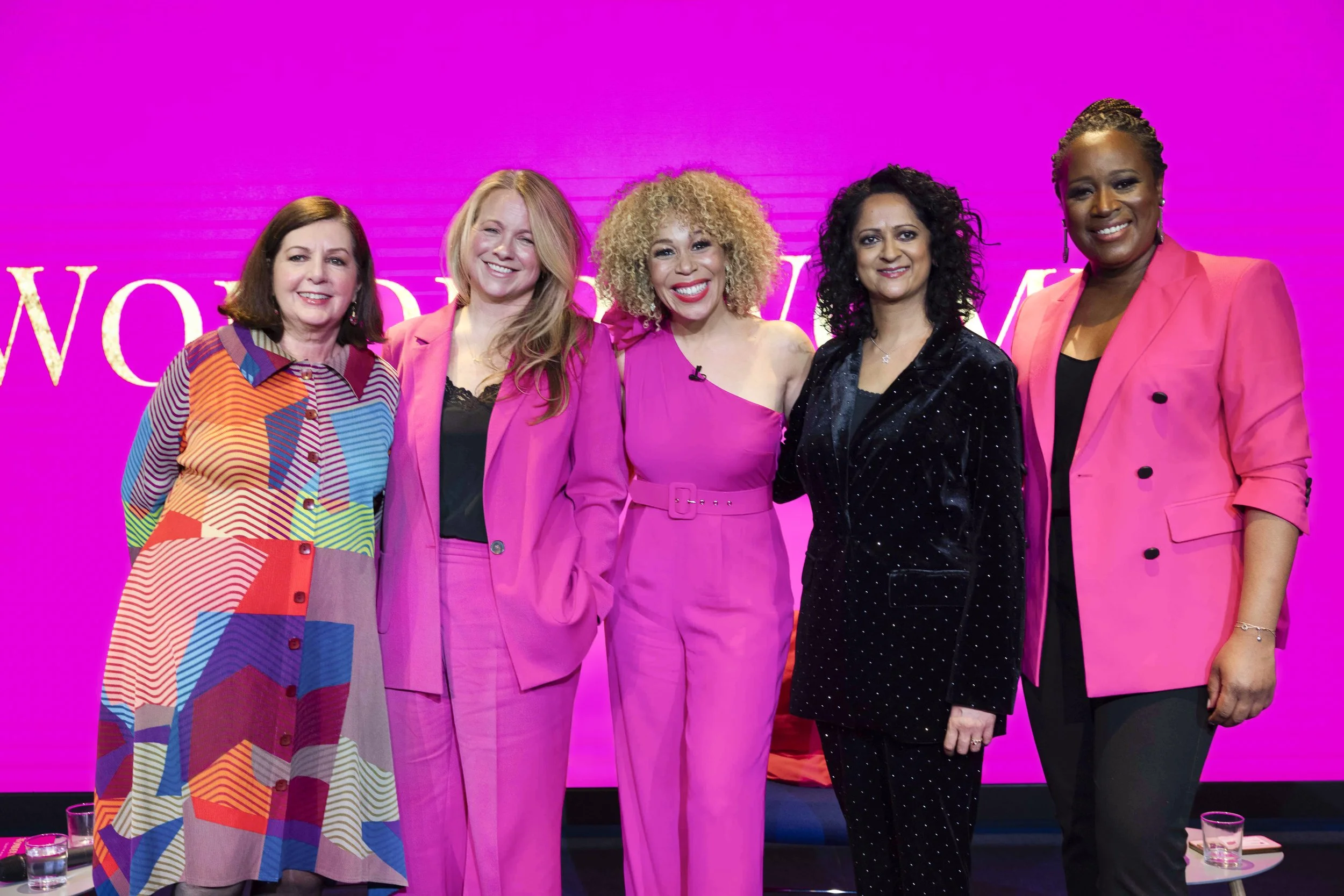 Group of five women standing together in front of a pink neon backdrop, dressed in colorful and pink outfits, smiling at the camera.