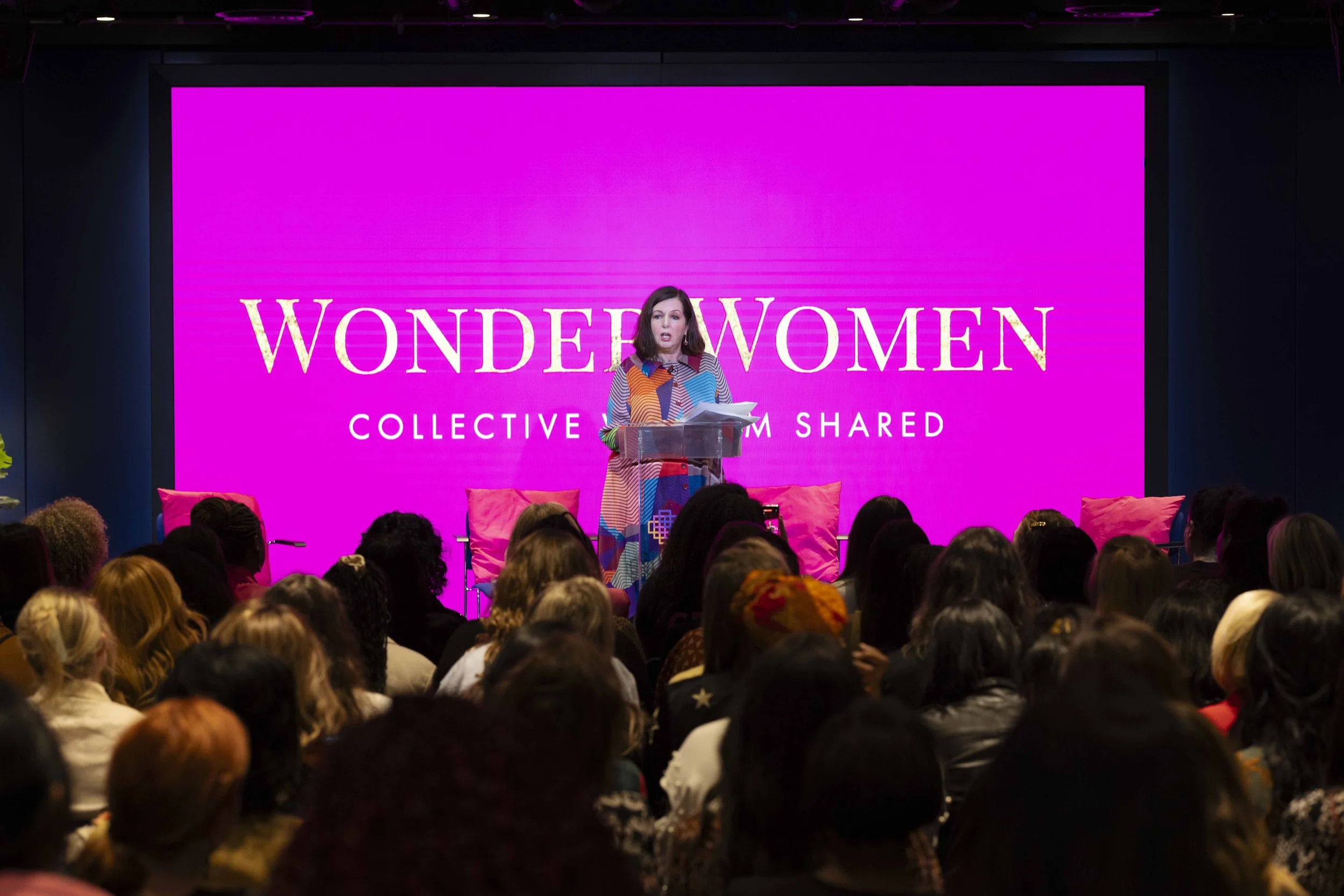 A woman is speaking at a podium in front of a large pink digital screen that reads 'WONDER WOMEN COLLECTIVE' in a room filled with a seated crowd.