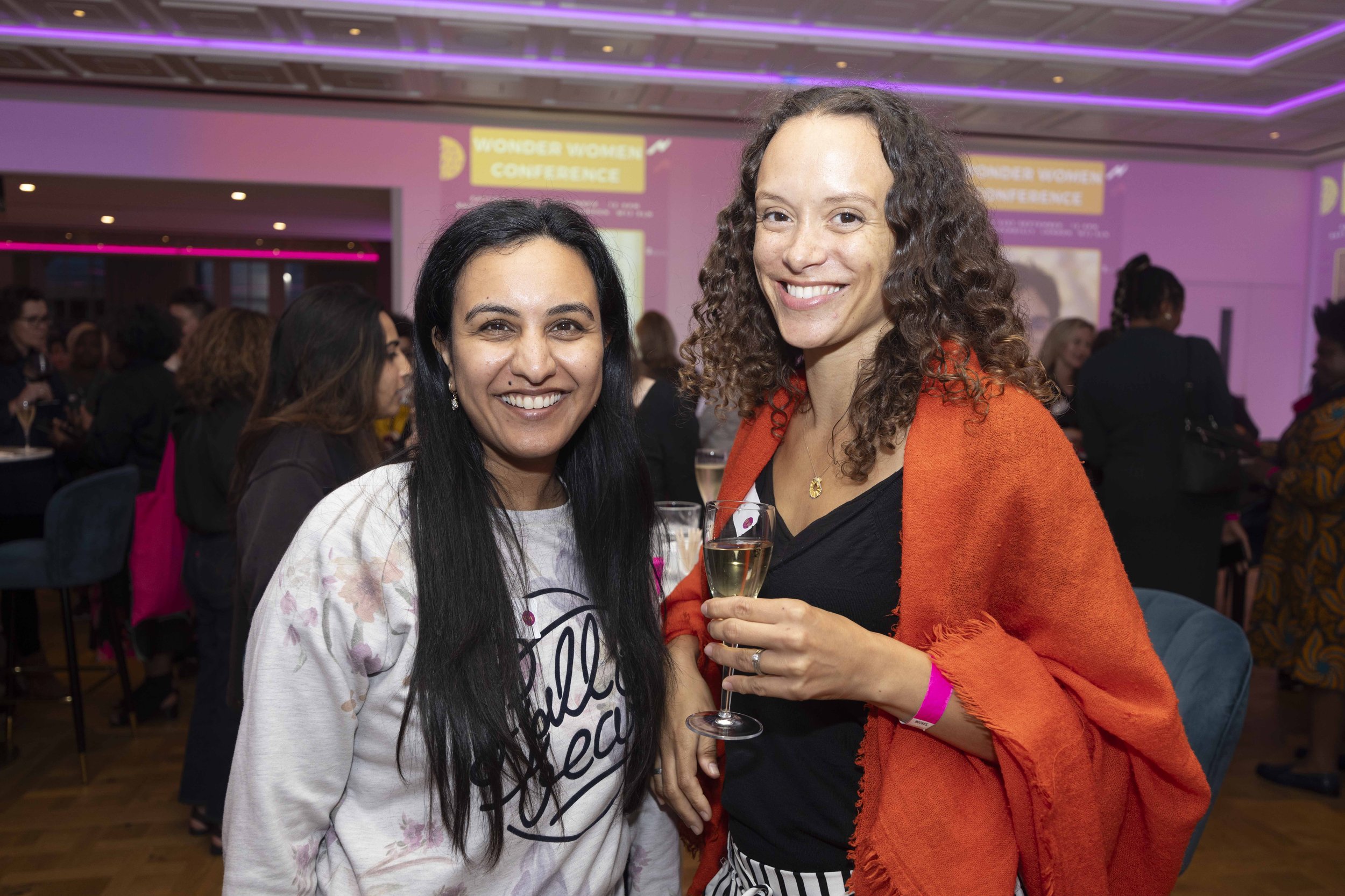 Two women smiling at an event, one holding a glass of white wine, with a pink and purple-lit conference room in the background.