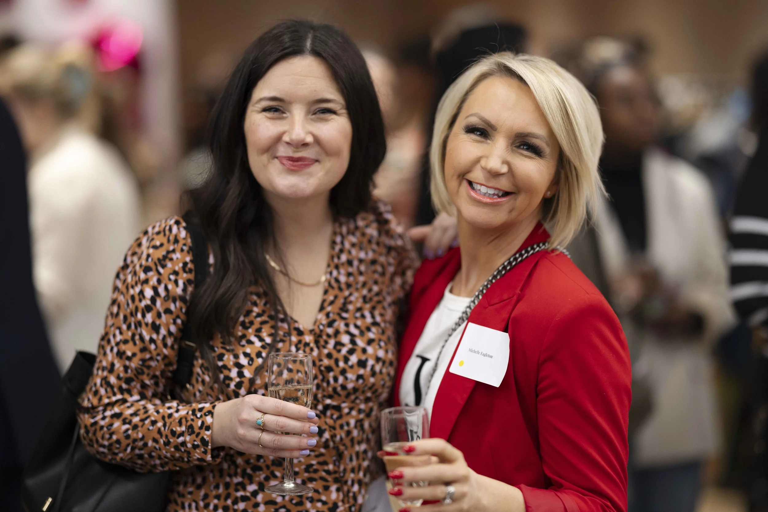 Two women smiling at a social event, holding glasses of champagne, with a blurred background of other attendees.