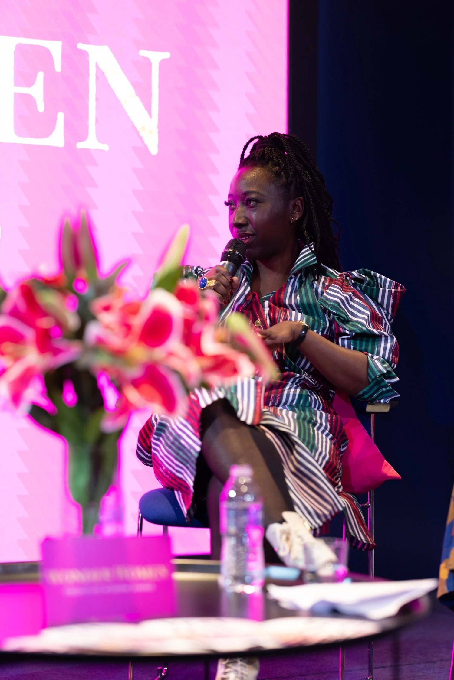 Woman in a colorful striped outfit speaking into a microphone on a stage with a pink background, foreground blurred with pink flowers and a table with a water bottle.