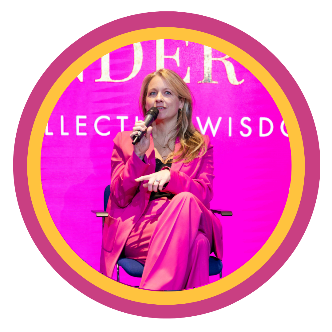 A woman in a pink suit sitting on a stage with a pink background, holding a microphone, during a speaking event.