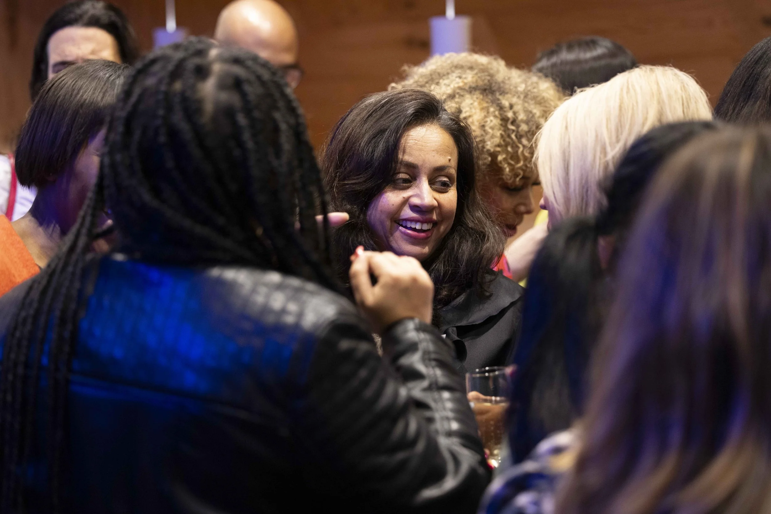 A group of people engaged in a lively conversation at an indoor gathering, with a woman smiling prominently in the center.