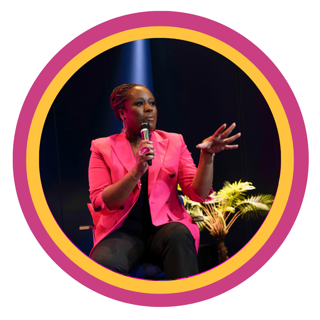 A woman in a pink blazer is speaking into a microphone during a talk or presentation, seated on a chair, with a dark background and a small plant nearby.