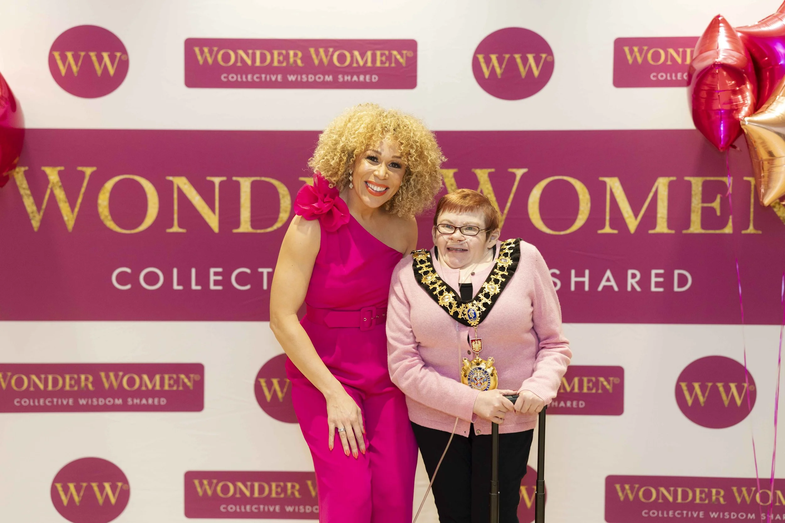 Two women standing together at a Wonder Women event, smiling and wearing medals, with balloons on the side and a pink backdrop featuring the Wonder Women logo and text.