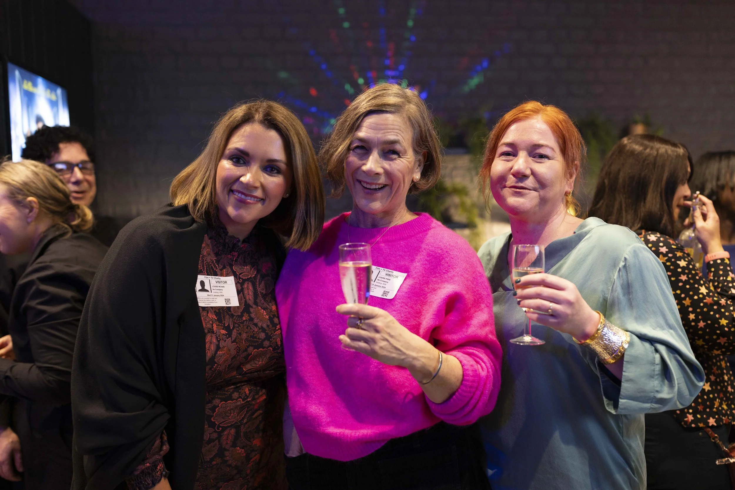 Three women smiling and holding glasses at a social event, with people and festive lighting in the background.