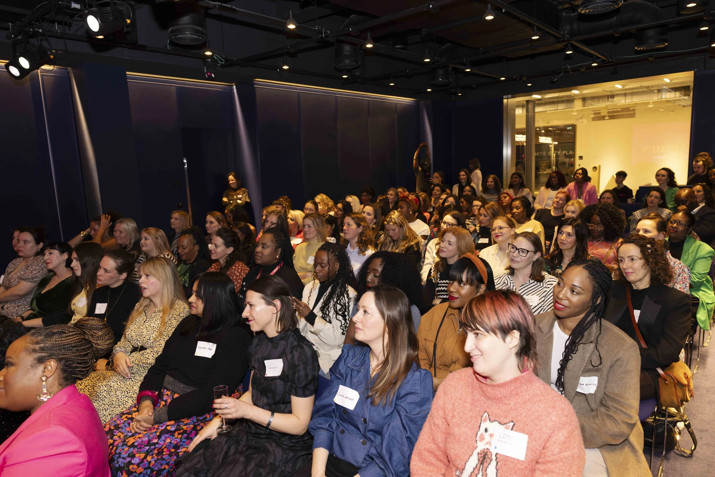 A large group of women attending a conference or seminar, seated and listening attentively in a dimly lit room with blue walls and ceiling lights.