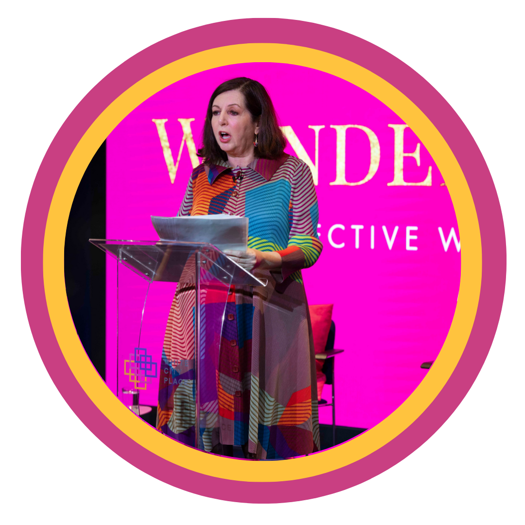 Woman in multi-colored dress speaking at a podium with a pink background displaying the word "WONDER" and partly visible text, possibly about an event or conference.