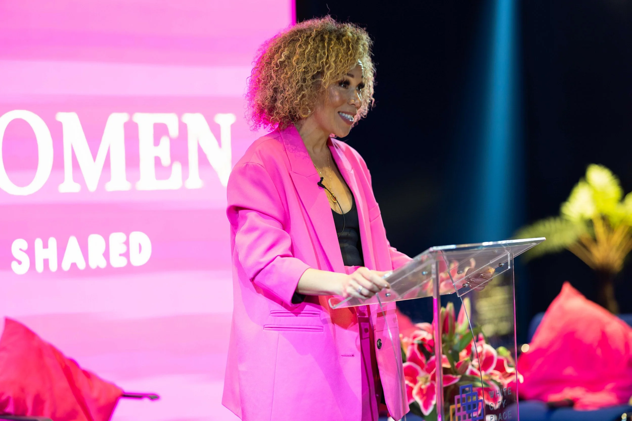 Woman in pink blazer speaking at a podium with a pink background featuring the word 'WOMEN.'