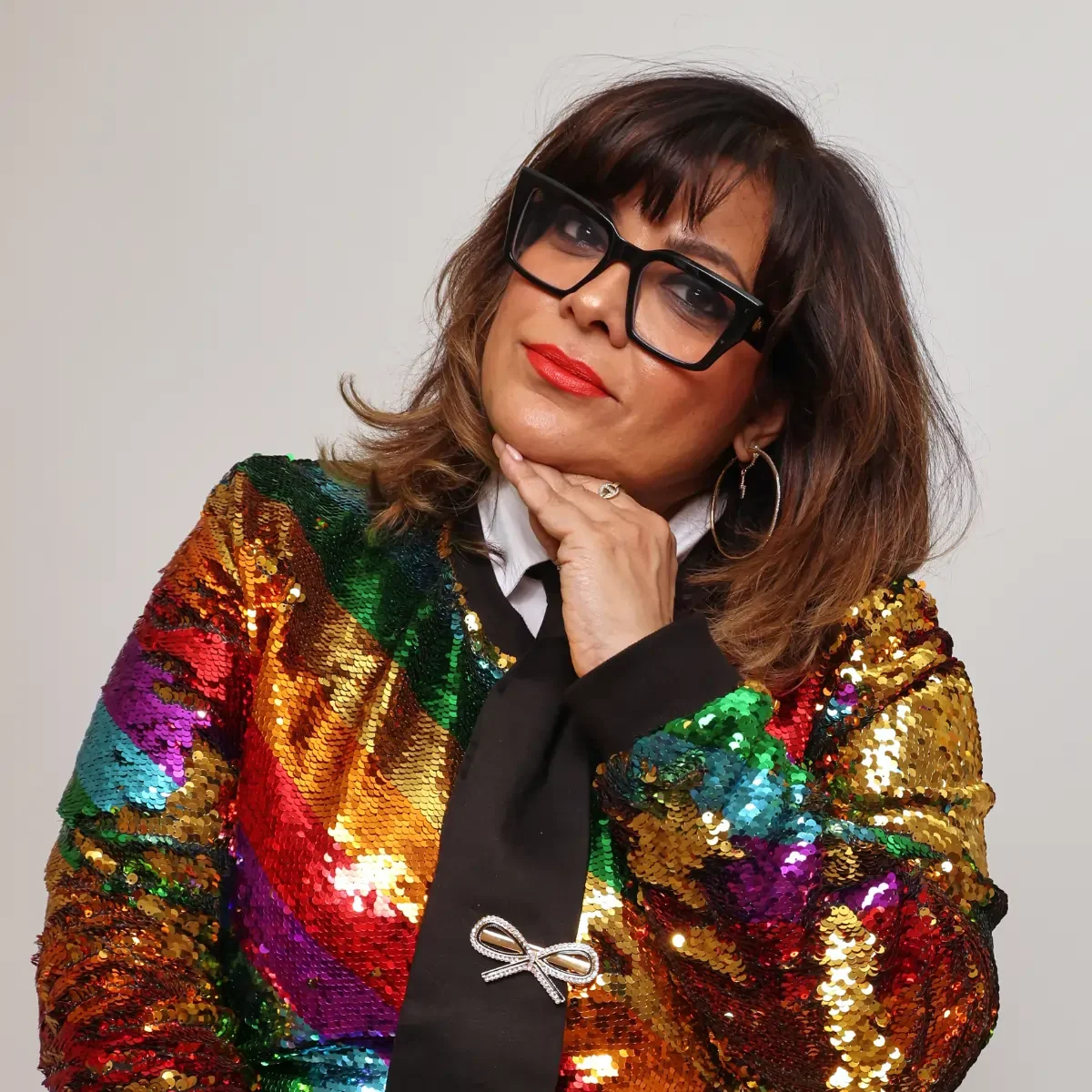 An asian woman wearing glasses and a multi-coloured, sequinned jacket poses confidently.