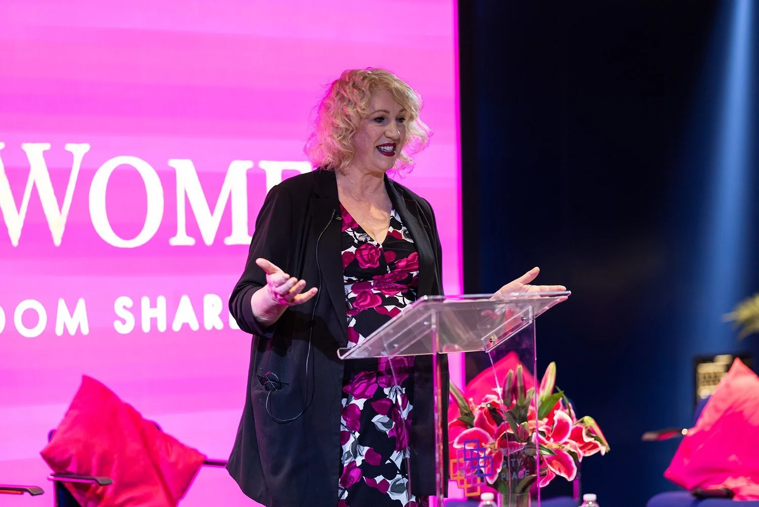 Woman speaking at a podium with a pink background screen and flower decorations.