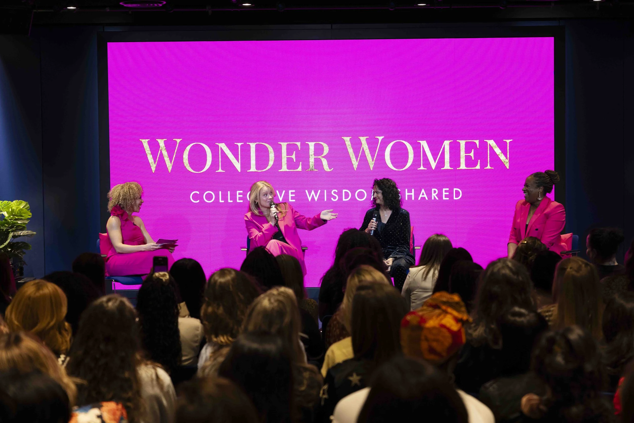 Four women in bright pink and black outfits sitting on stage in front of an audience, engaging in a panel discussion at an event called Wonder Women, with a large pink screen behind them displaying the event's title.