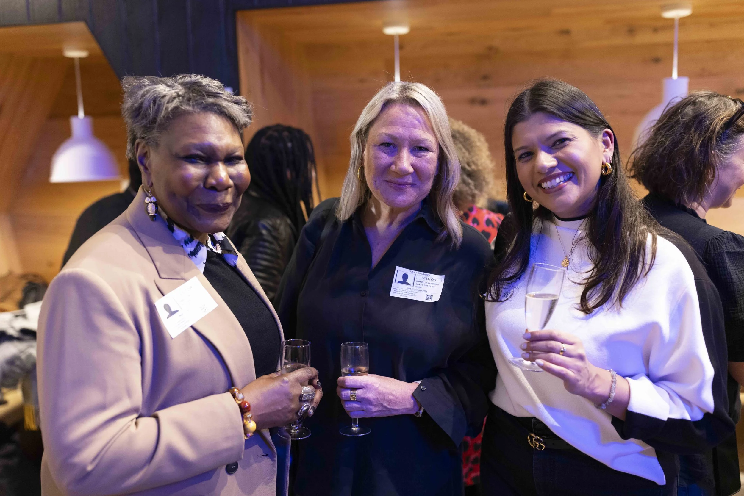 Three women smiling, holding glasses at a social event, wearing visitor name tags.