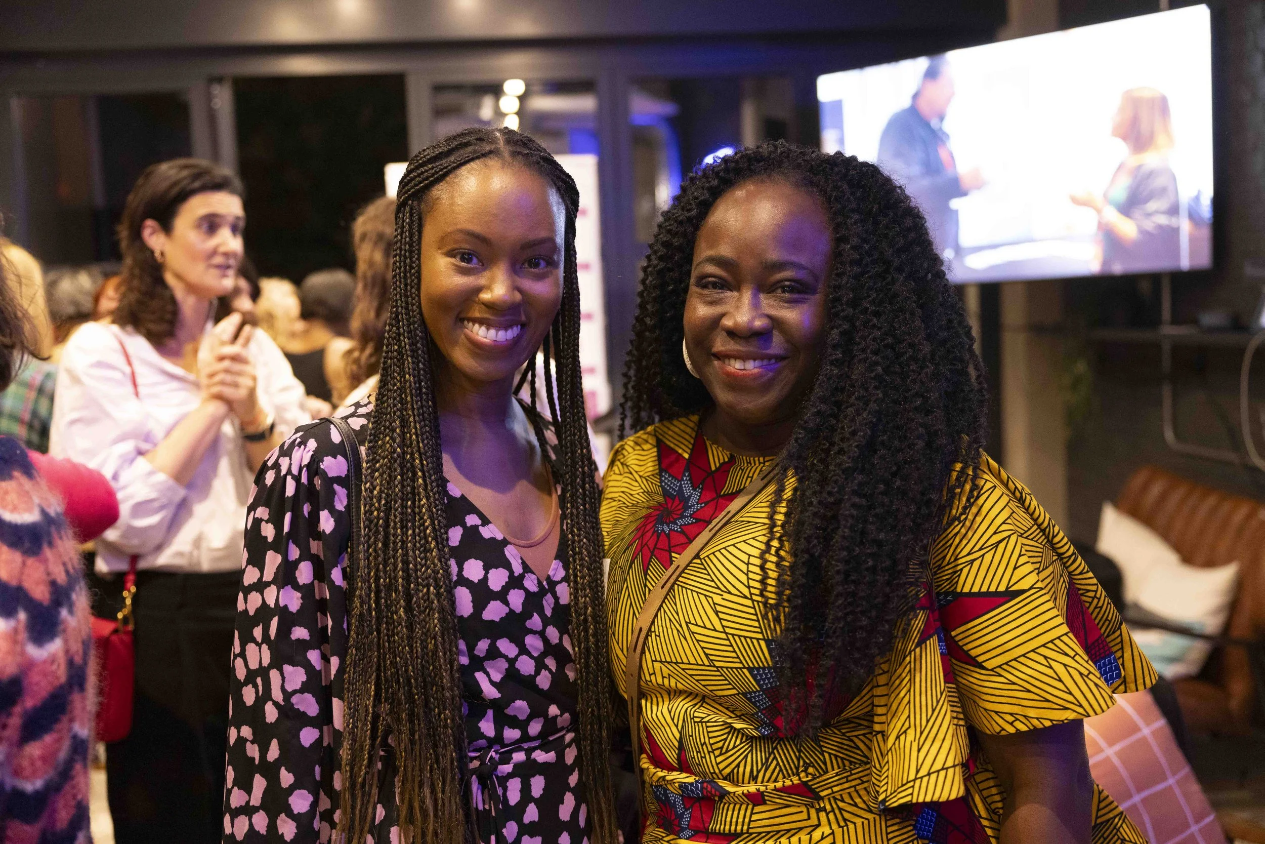 Two women smiling at a social event with other guests in the background. One woman is wearing a pink and black patterned dress, the other a brightly colored yellow and red patterned dress.