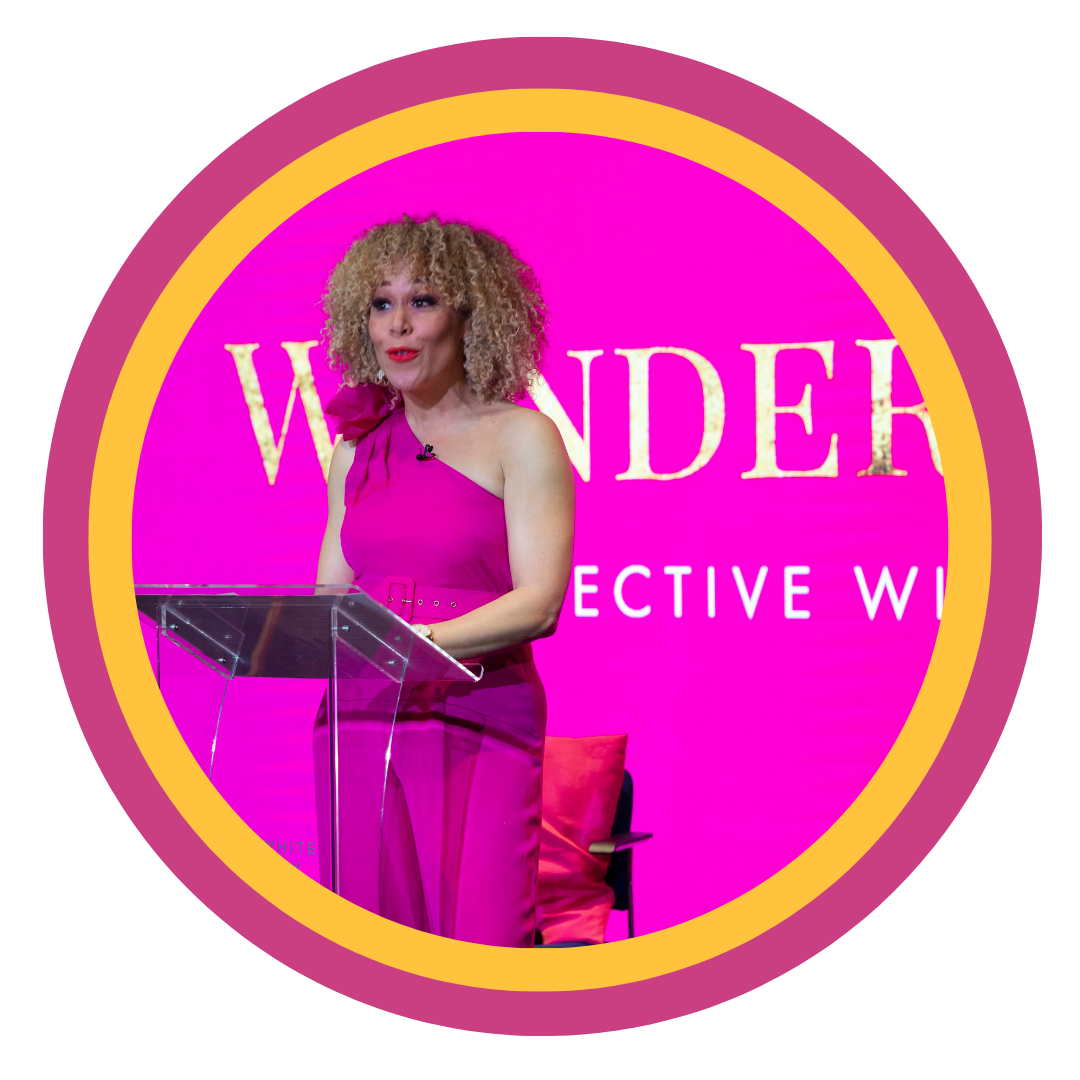 Woman with curly blonde hair wearing a pink one-shoulder dress speaking at a podium with a pink background that says "WANDER" and "COLLECTIVE W".