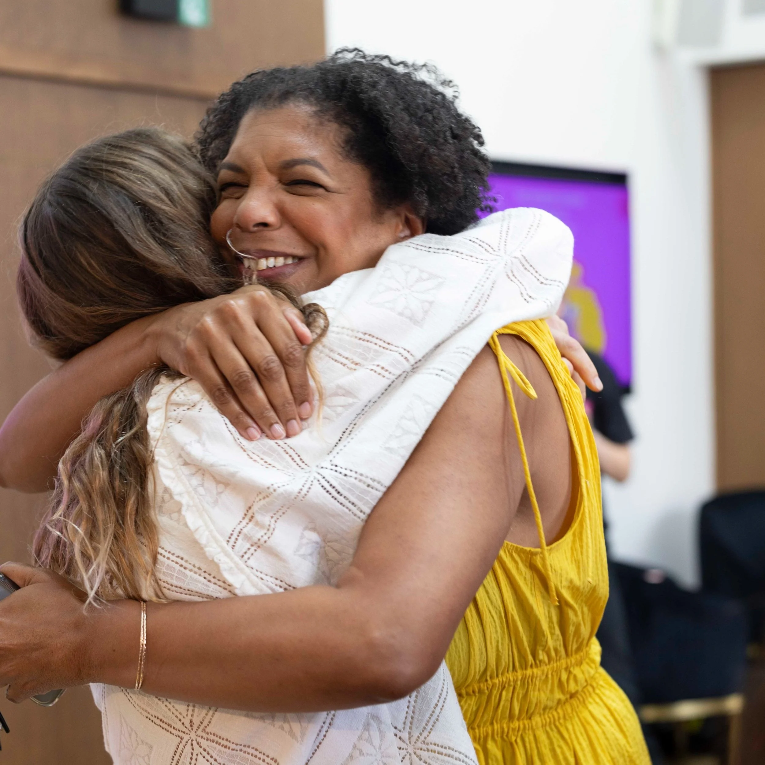 Two women hugging each other warmly, one wearing a white embroidered top and the other in a yellow dress, both smiling.