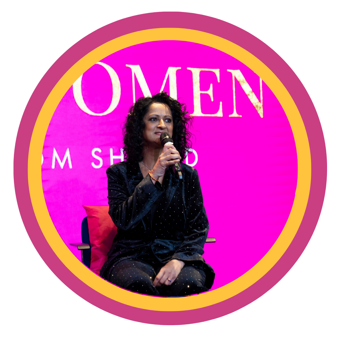 A woman sitting on a chair with a pink pillow, speaking into a microphone at an event with a pink background that says 'Women' and other words.
