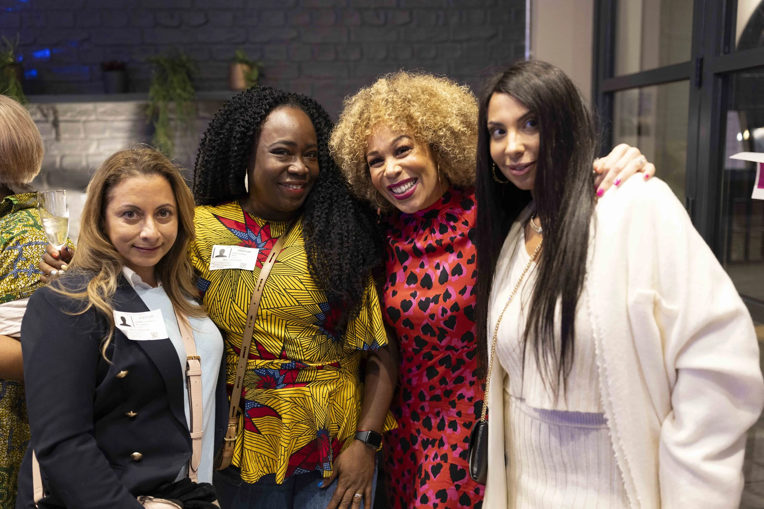 A group of four women smiling at a social event, wearing name badges. They are dressed in colorful and stylish outfits, standing indoors with a brick wall and decorations in the background.