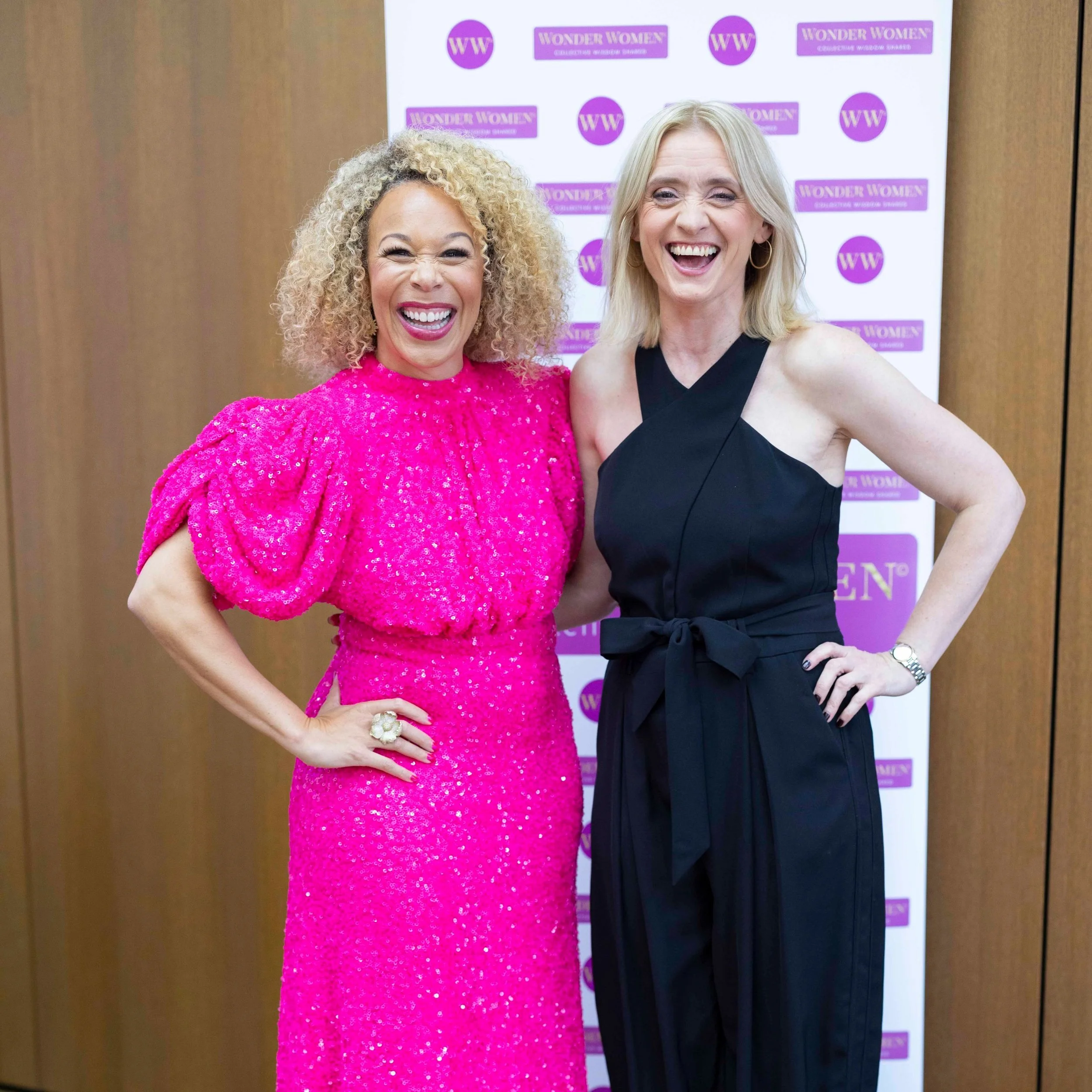Two women standing together and smiling, in front of a purple backdrop that reads 'Wonder Women'. The woman on the left wears a bright pink, sequined dress with puffed sleeves, and the woman on the right wears a sleeveless black jumpsuit with a tied waist. Both women have light-colored hair and are wearing jewelry.