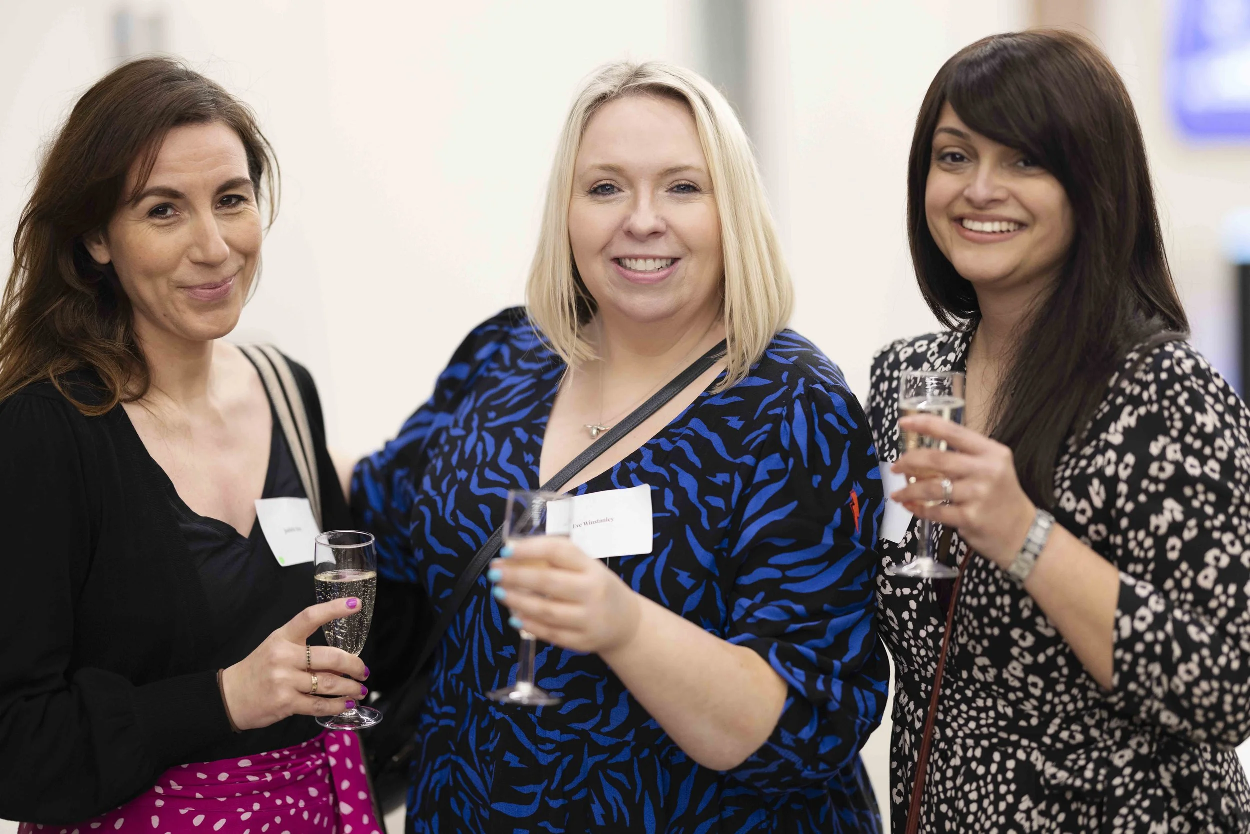 Three women at a social event holding glasses of champagne, smiling, standing in front of a neutral background.