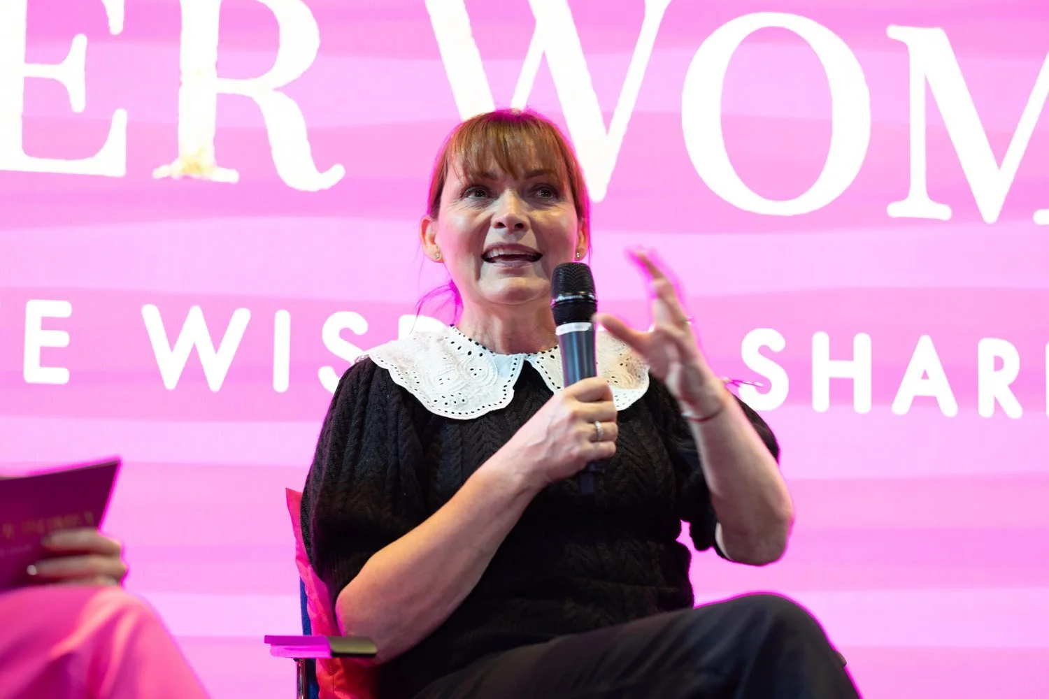 A woman holding a microphone while speaking on a stage with a pink background. She wears a black outfit with a white lace collar. Partial text behind her is visible, reading "WISE" and "SHAR".