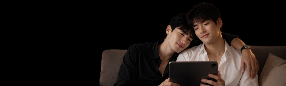 Two young men sitting close together on a couch, looking at and smiling at a tablet device, with one man's arm around the other's shoulder.