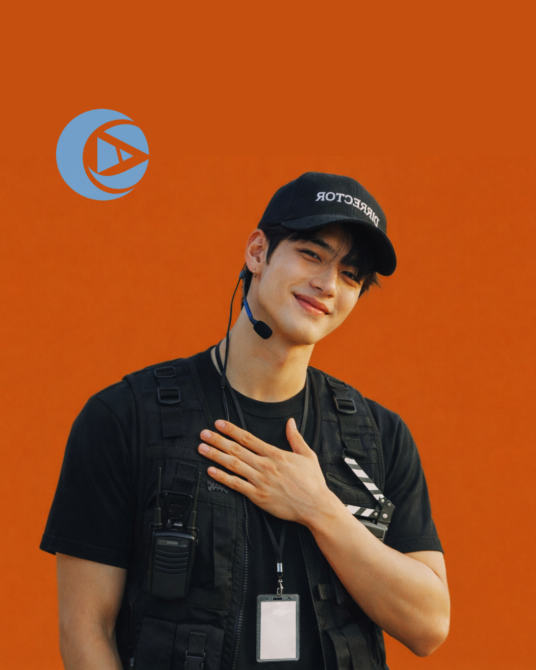 Young man wearing a black 'Director' cap, black t-shirt, and a black tactical vest with a walkie-talkie, badge, and a headset. He is smiling, with his right hand over his chest, against an orange background with a blue logo in the top left corner.