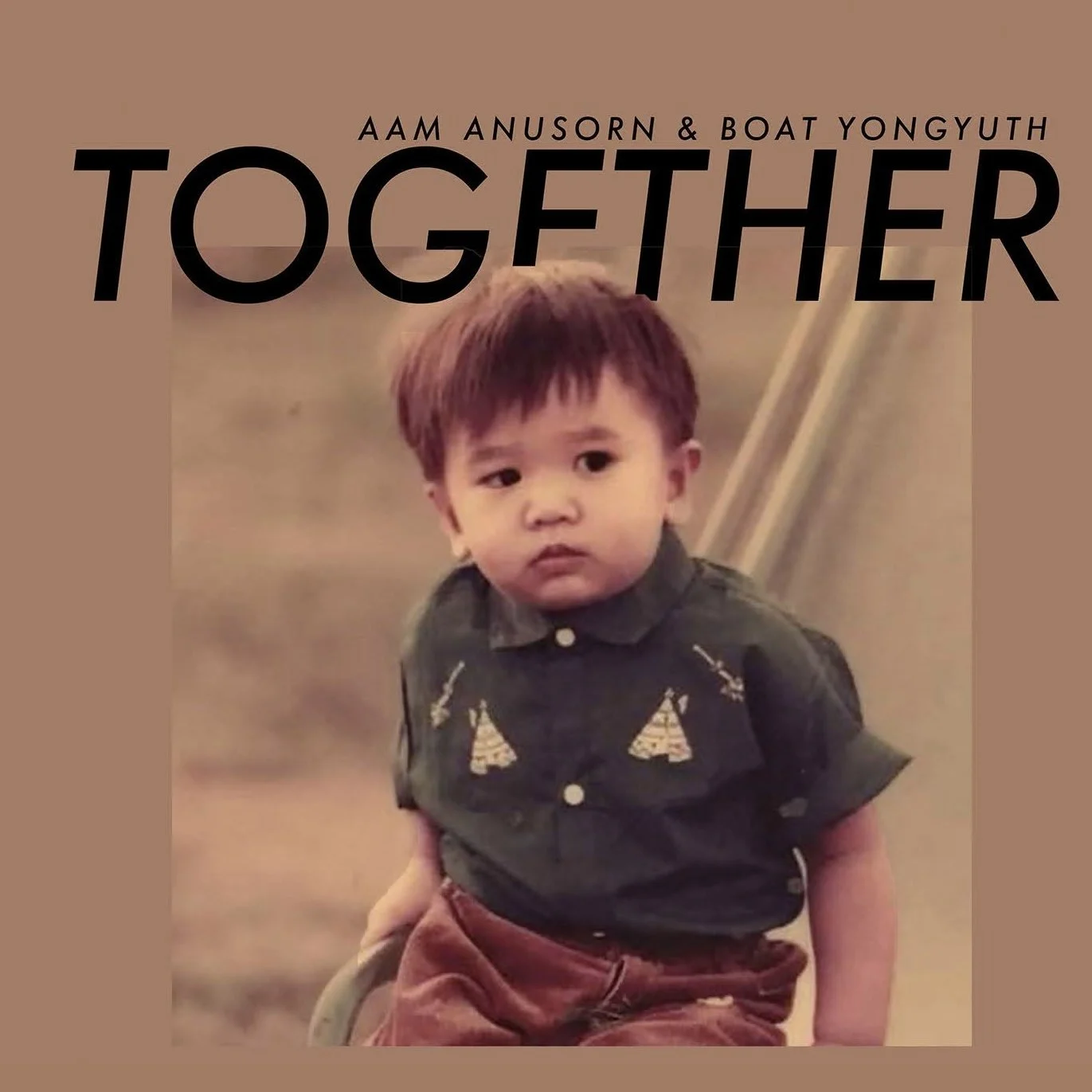 A young boy with brown hair wearing a black shirt with trees embroidered on it, sitting or leaning on a chair, looking serious.