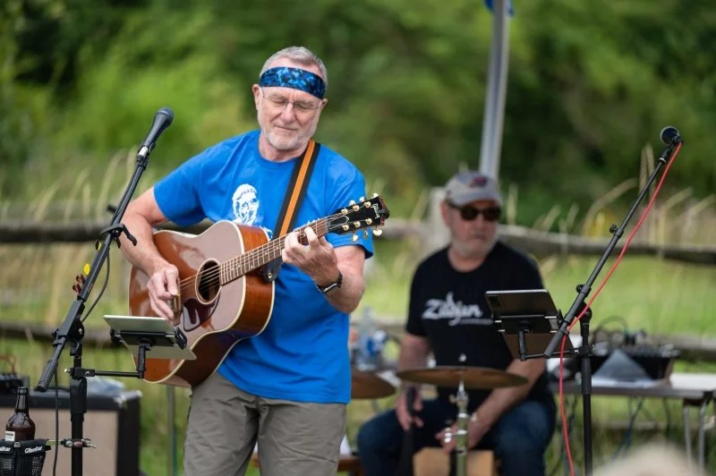 A man playing an acoustic guitar in front of a drummer in an open air setting