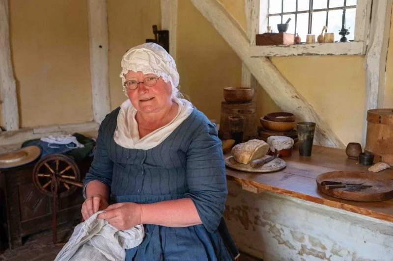 A woman dressed in a blue dress and white cap sits in a wood framed cottage next to a spinning wheel and a wooden table that has wooden bowls on it