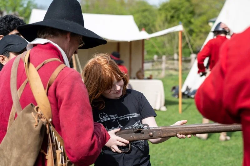 A young boy holds a replica English Civil War gun while talking to costumed re-enactors dressed as civil war soldiers