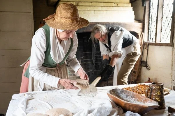A woman in 18th century costume places a ball of risen dough onto a wooden tool that's held by a man waiting to put the loaves into the oven in an 18th century cottage