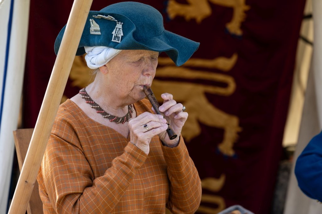 An older lady dressed in simple medieval costume plays a small wooden recorder