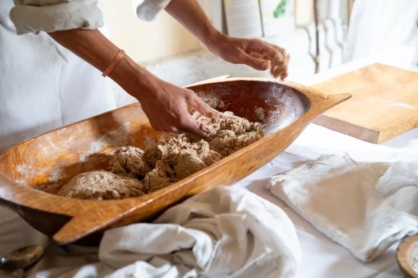 Hands mix together flour, yeast, salt and warm water in a large shallow oval wooden bowl known as a trow