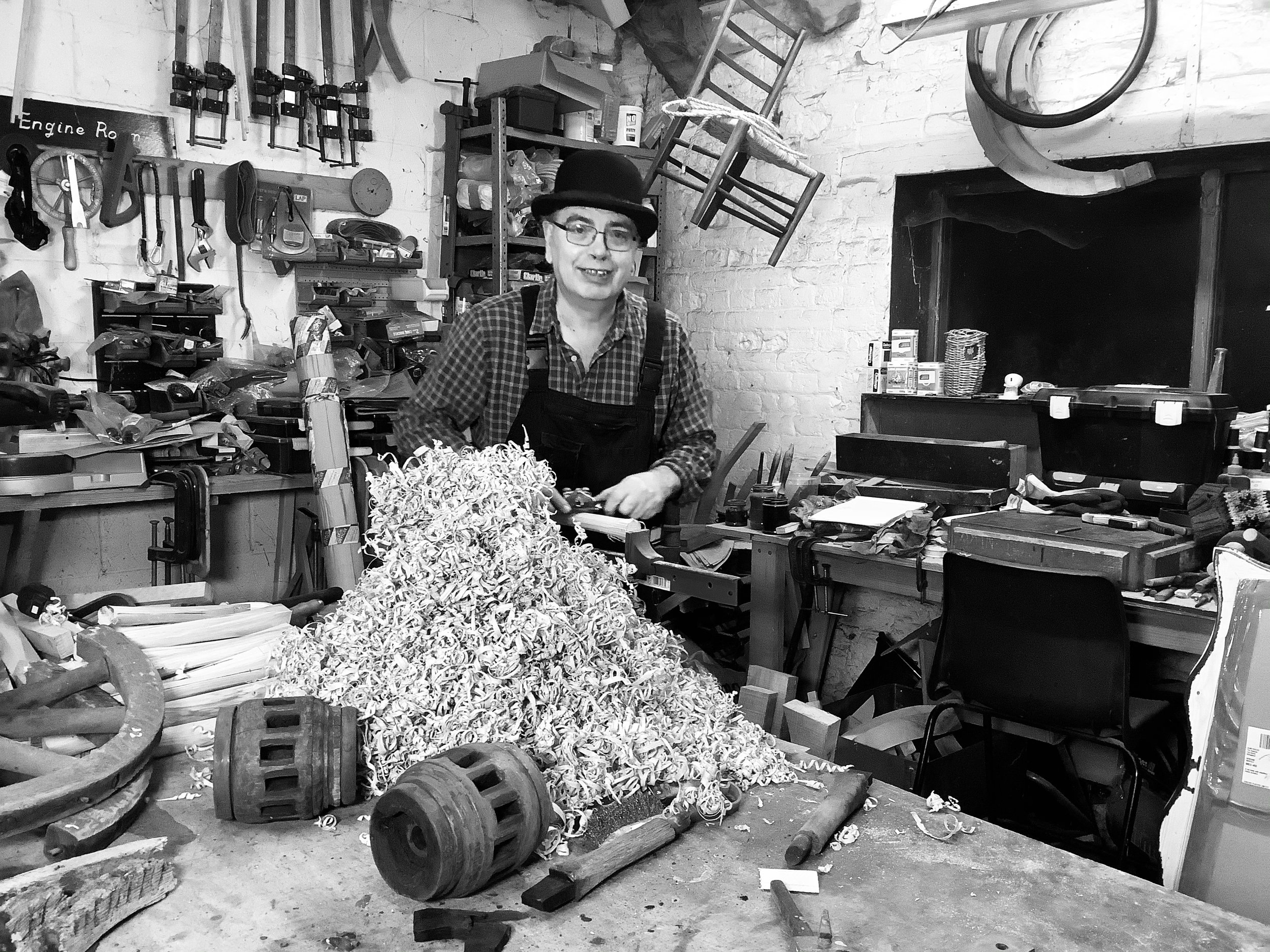 A smiling man wearing a bowler hat sits in a busy workshop surrounded by tools.