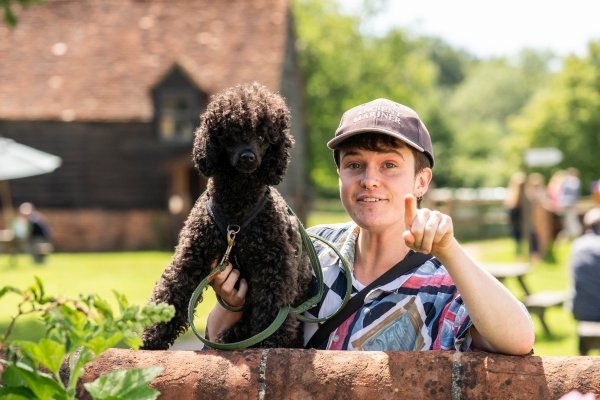 A young person is standing behind a brick garden wall holding a black small poodle, smiling and pointing at the camera.