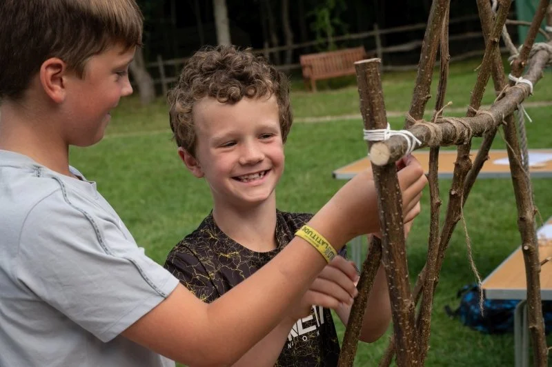 Two smiling boys making a structure out of sticks and string