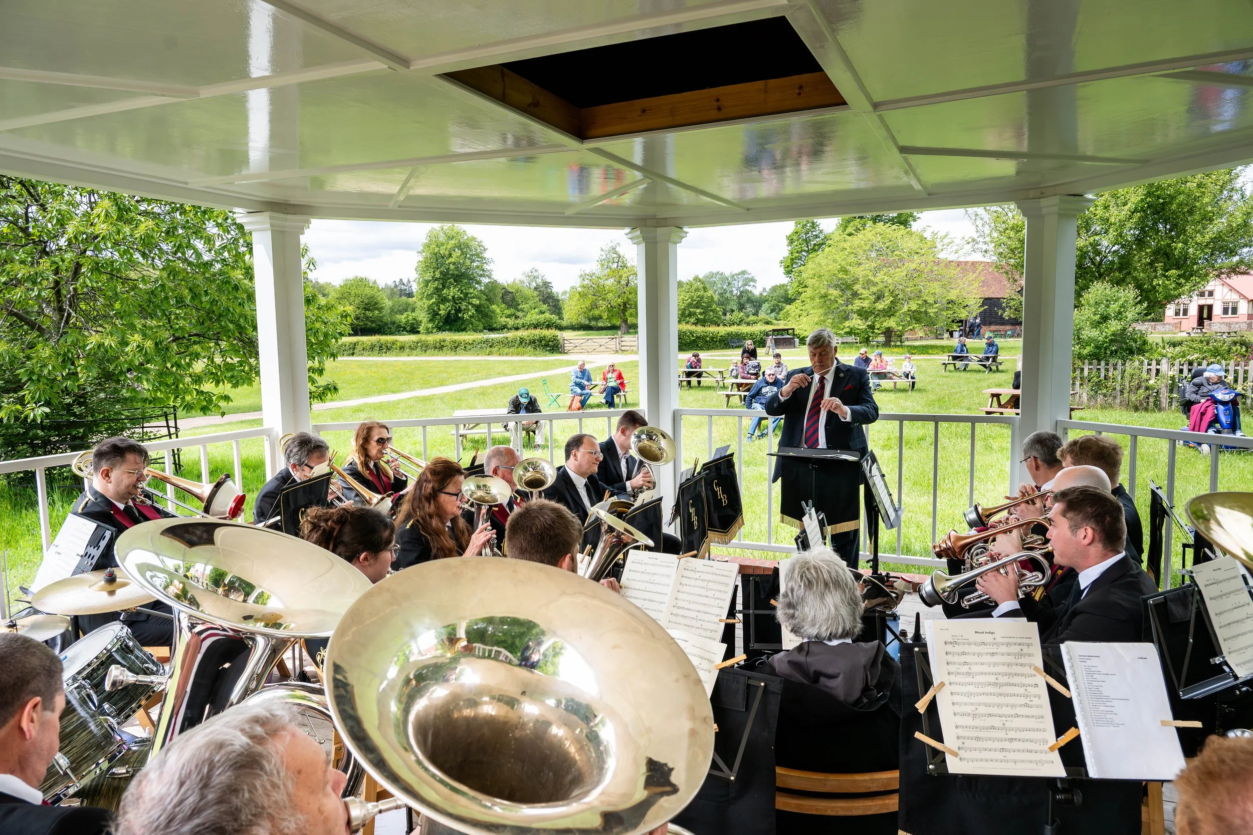 A brass band playing on a bandstand surrounded by green grass and trees with people watching from picnic tables