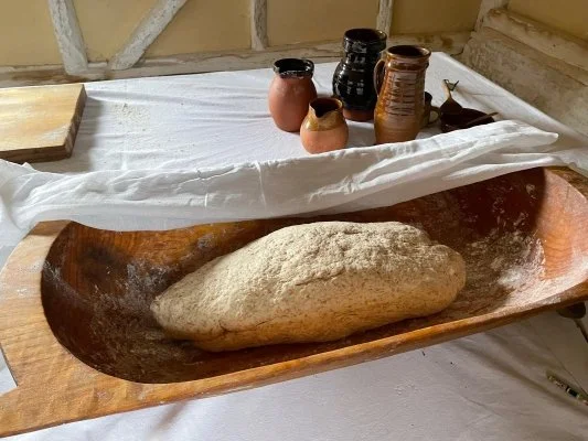 A large oval wooden bowl called a trow containing a piece of dough sits on a table in an 18th century cottage.