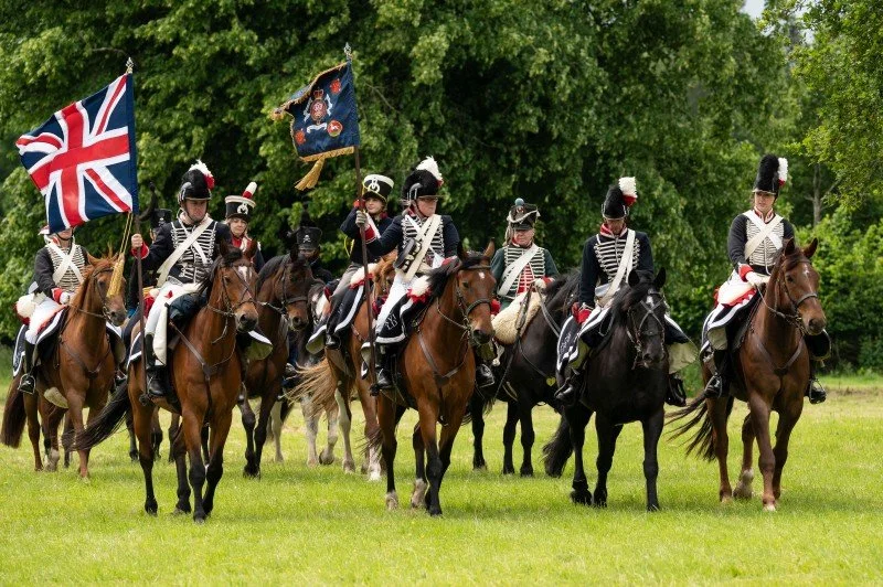 A group of re-enactors dressed in the uniform of the Napoleonic Wars ride horses and carry  British flags