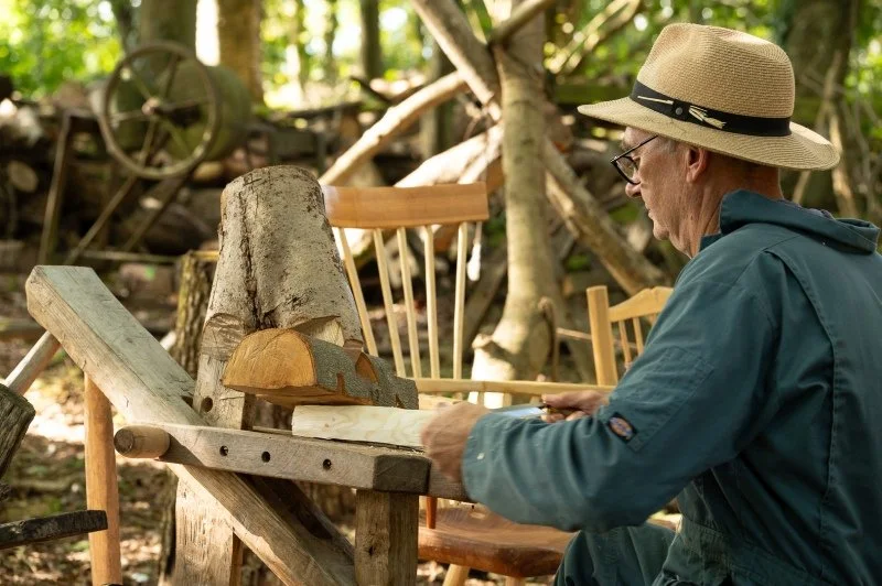 A man using wooden tools to craft chair legs out of logs sits in a woodland