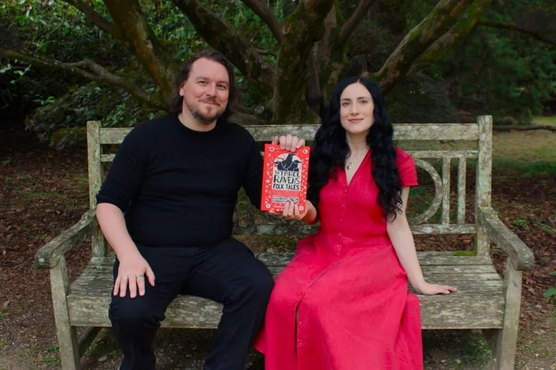 A man and a woman sit on a bench holding a Three Ravens Folk Tales book