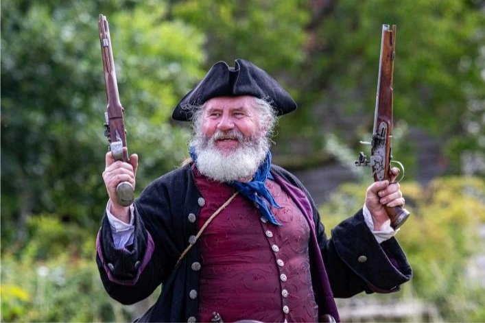 A smiling man dressed in a three cornered hat and 18th century clothing raises two vintage handguns